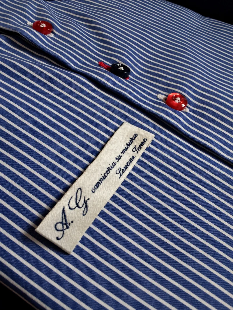 Camicia a righe blu e bianche con bottoni rossi e blu ed etichetta della camiceria Antonio Gigliotti.