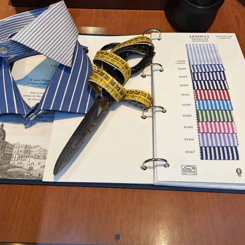 Forbici da sartoria e metro giallo su un campionario di tessuti, accanto a un collo di camicia a righe.
